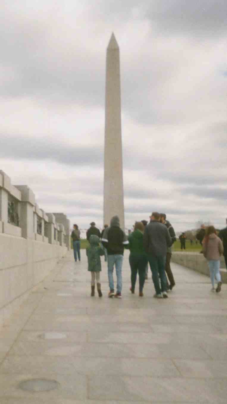 About a dozen visitors are seen walking on a stone pathway toward the Washington Monument, which, visible in the background, rises to more than 555 feet.