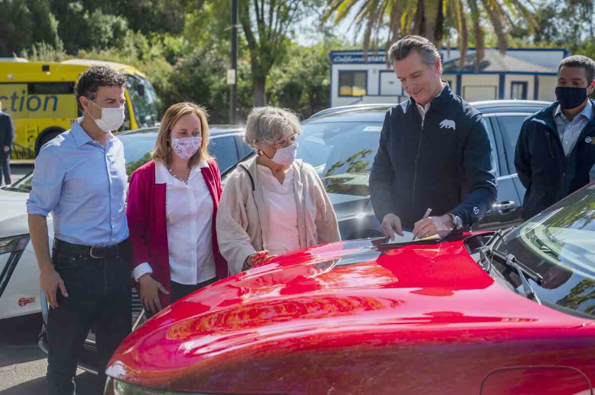 Gov. Gavin Newsom unterzeichnet im Freien eine Executive Order auf der Motorhaube eines roten Autos, während vier Personen zuschauen.