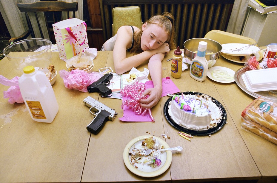 a young woman lays her head on a table filled with food and toy guns after a party.