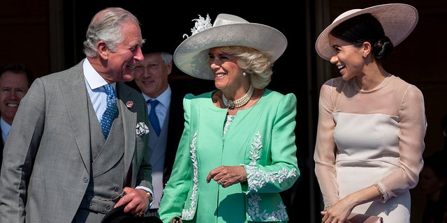 King Charles in einem grauen mehrteiligen Anzug lächelt Meghan Markle in einem errötenden Kleid und Hut an, in der Mitte lächelt Camilla in einem hellgrünen Kleid und einem weißen Hut und sieht Charles an