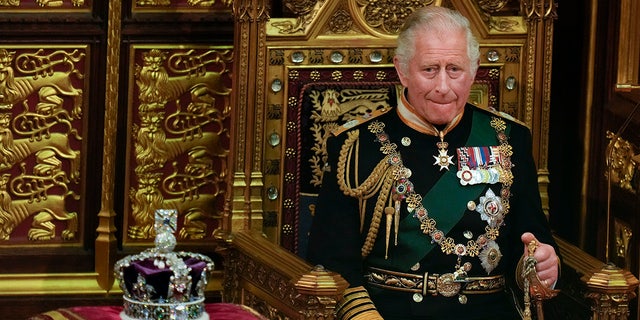 König Charles im grünen Anzug, geschmückt mit Orden und Ketten, sitzt auf einem königlichen Stuhl neben einer Krone