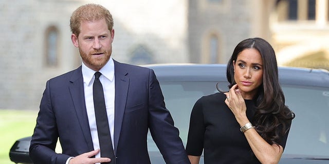 Prinz Harry in einem marineblauen Anzug und Krawatte hält Meghan Markles Hand, die nach dem Tod der Königin ein dunkles Kleid trägt