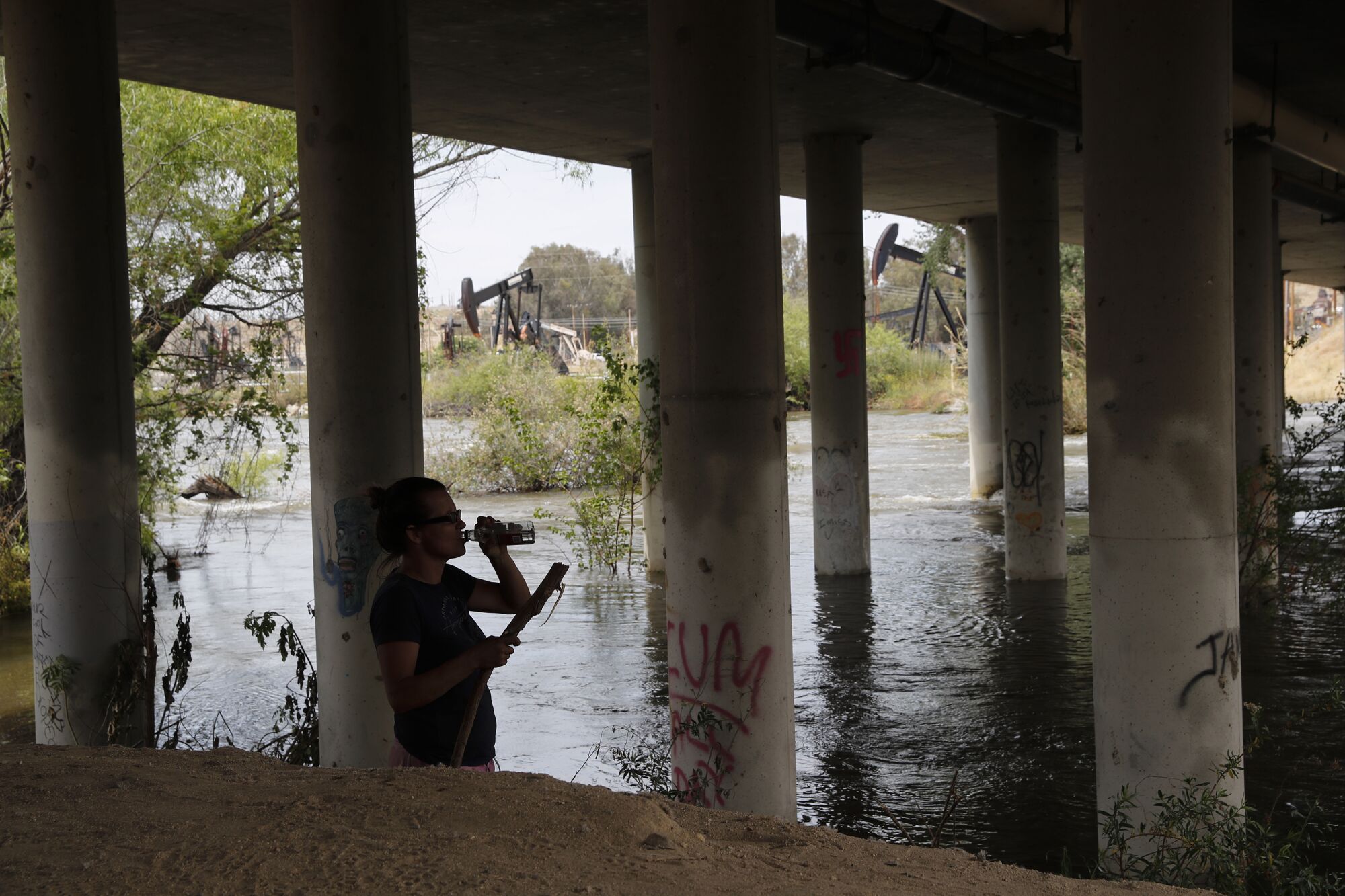 Eine Frau trinkt aus einer Flasche an einem Flussufer unter einer Brücke.