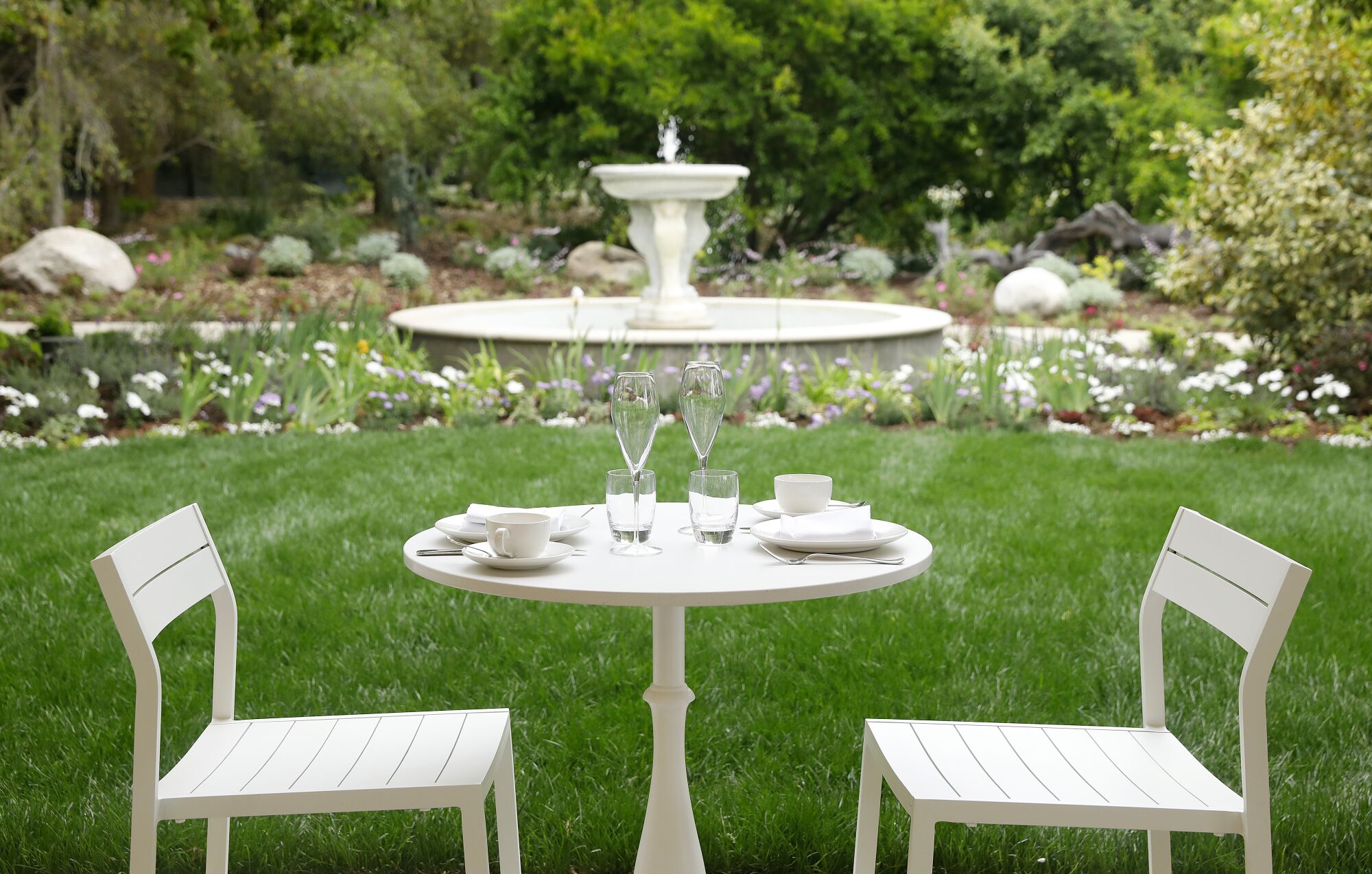 Ein weißer Tisch mit zwei Stühlen, dahinter ein Gartenbrunnen.