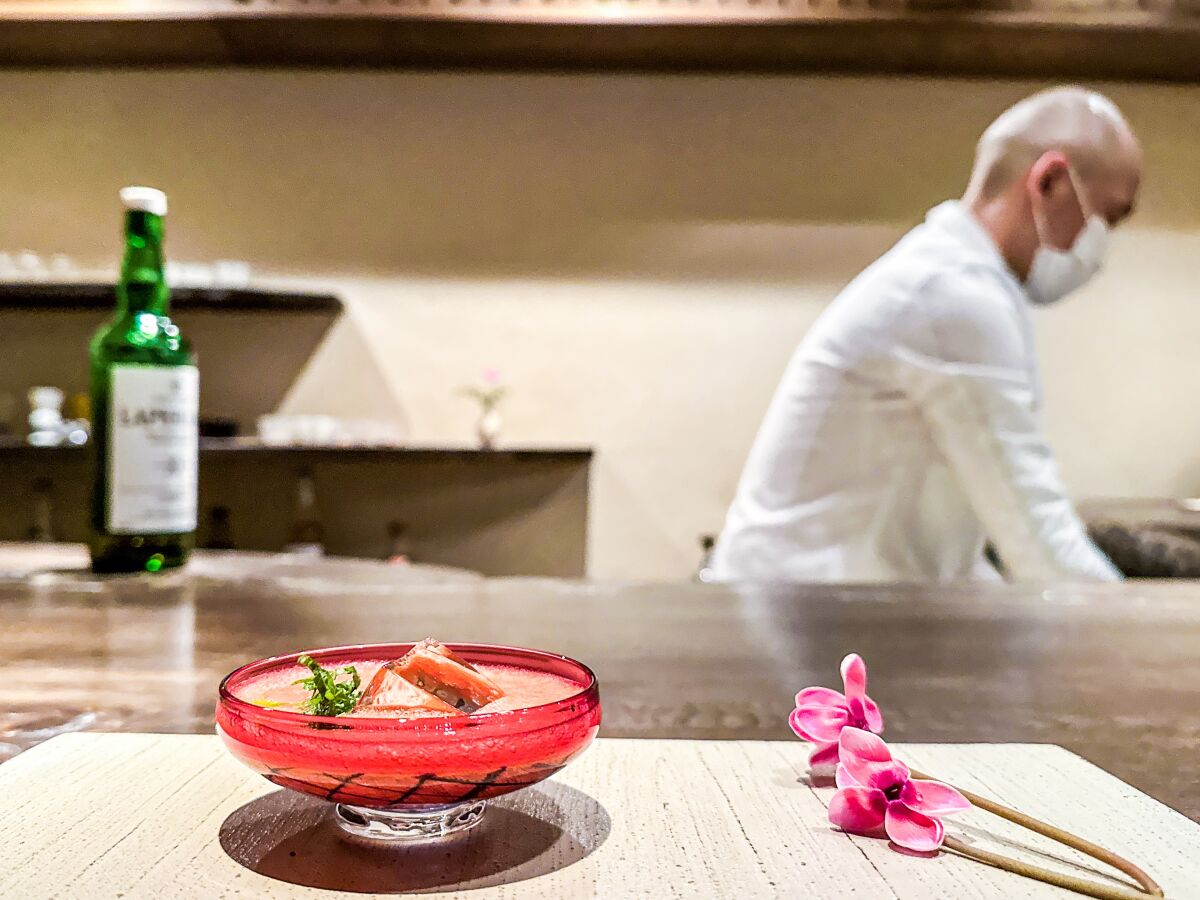Ein Cocktail in einem flachen Glas steht auf einer Theke, während im Hintergrund ein Koch arbeitet