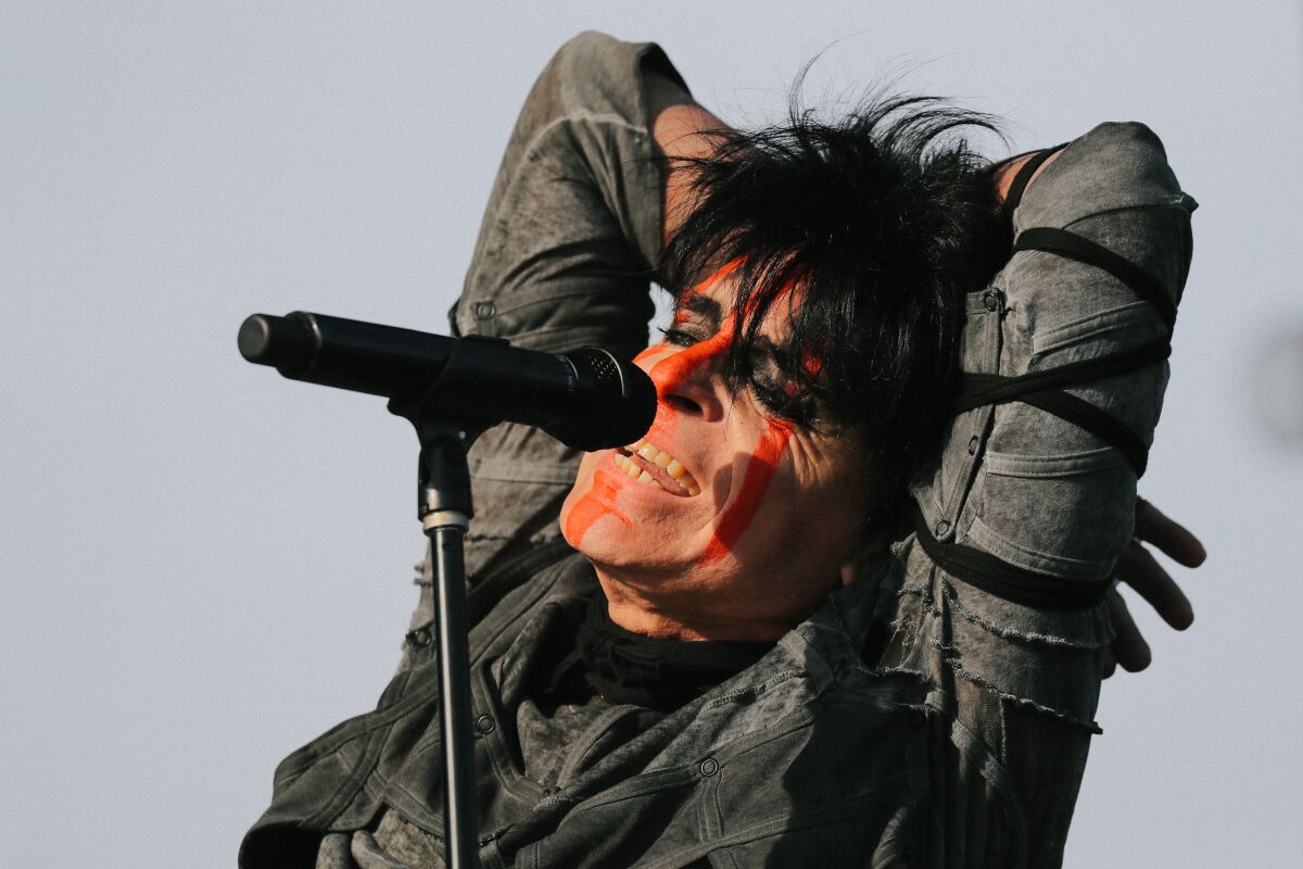 An einem Mikrofon steht ein Mann mit gefärbten schwarzen Haaren und strähnig orangefarbener Gesichtsbemalung, die Arme hinter dem Kopf verschränkt.