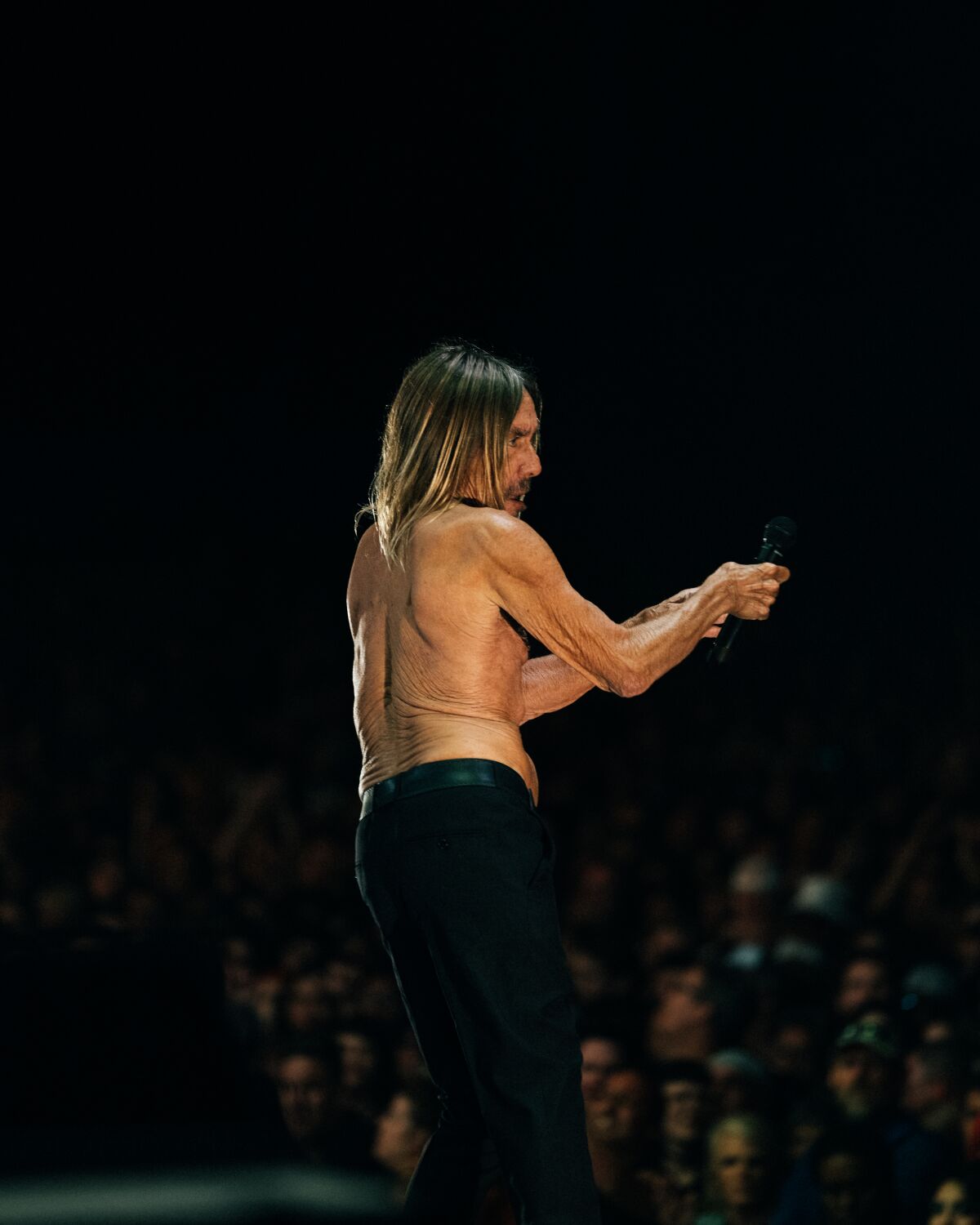 Ein Mann ohne Hemd und mit langen Haaren tritt vor Publikum auf der Bühne auf.