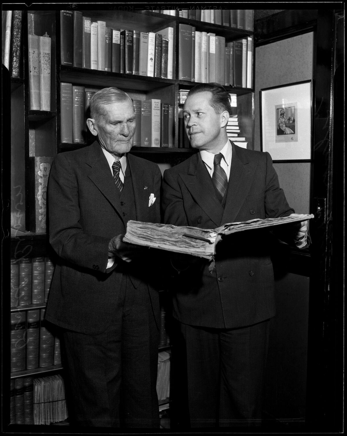 Auf einem Schwarz-Weiß-Foto stehen zwei Männer vor einem Bücherregal, einer hält ein großes aufgeschlagenes Buch. 