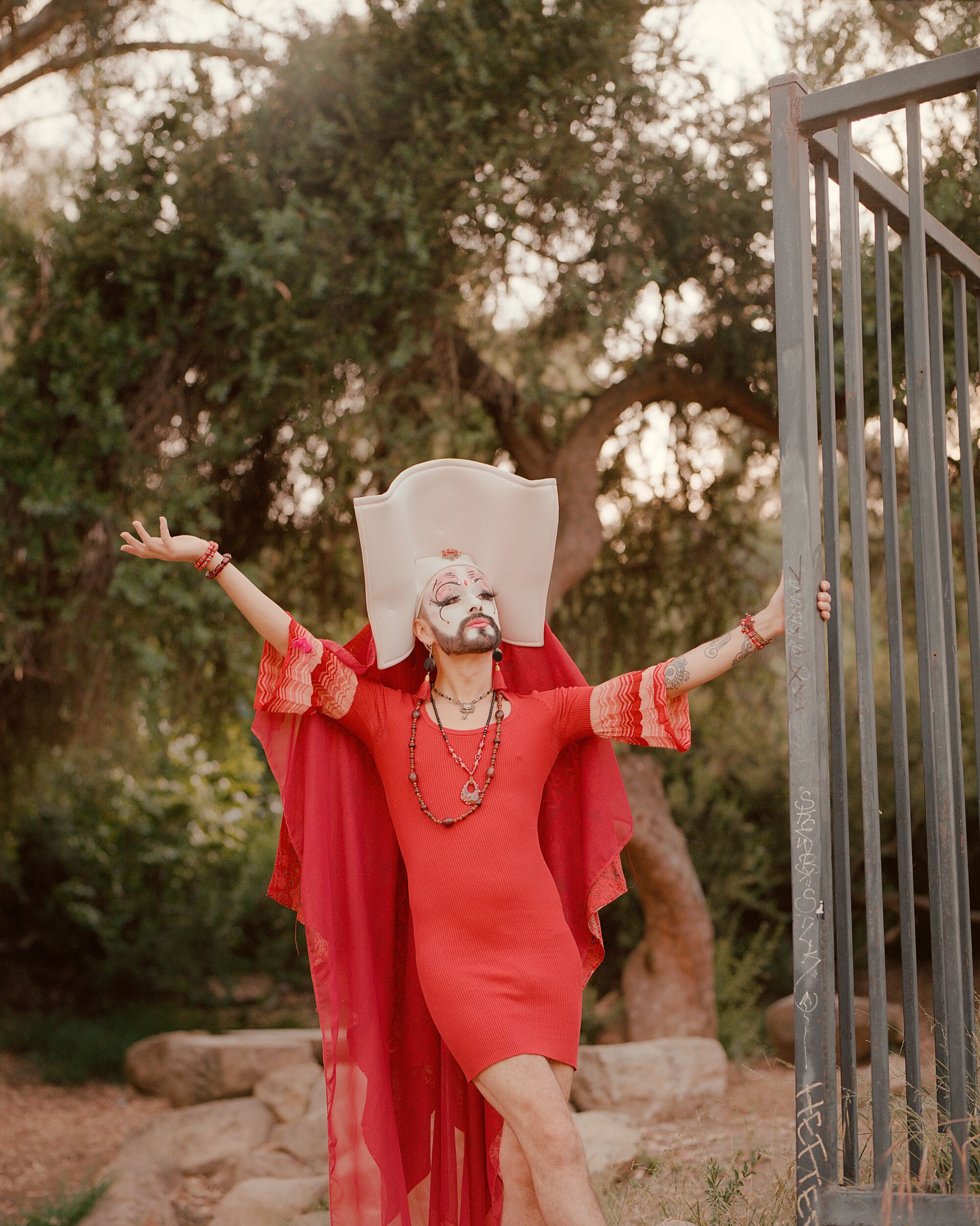 Eine Drag Queen in rotem Kleid, Bart, Gesichtsbemalung und kunstvollem Wimpole-Kopfschmuck posiert im Freien.