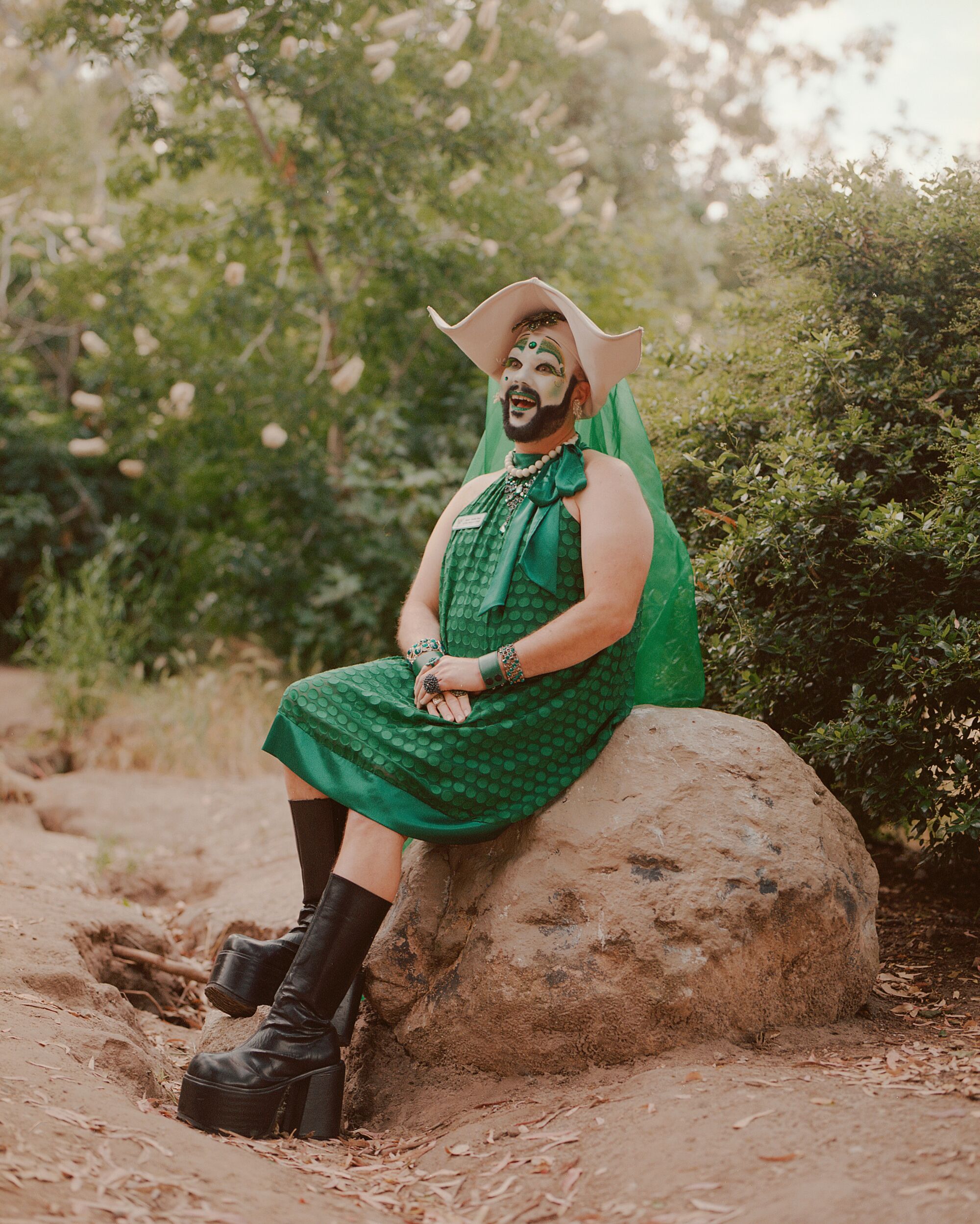 Eine Drag-Nonne in grünem Kleid und Stiefeln sitzt draußen auf einem Felsbrocken.