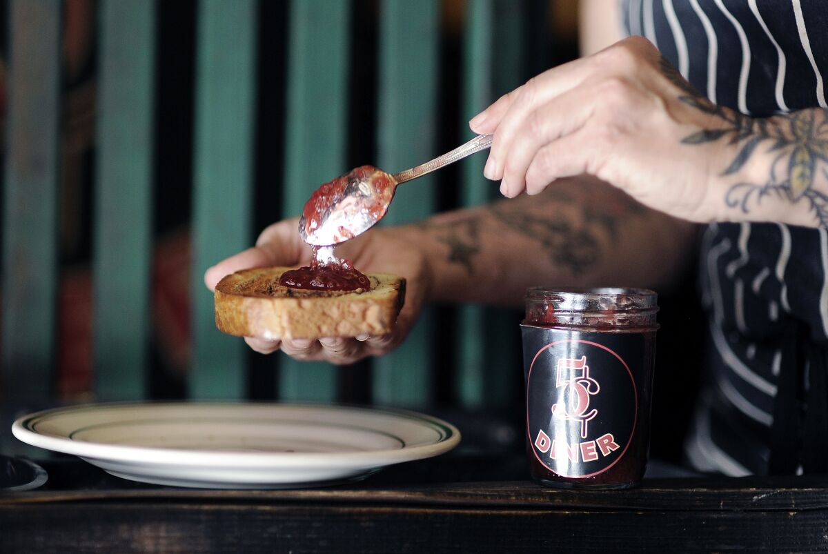 Eine Hand löffelt Erdbeermarmelade auf ein Stück Brot.  Vorne steht ein Glas mit Marmelade.