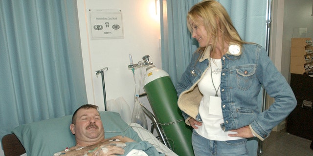 Bo Derek trägt eine Jeansjacke und Jeans mit passendem weißen Hemd und spricht mit einem Mann, der auf einem Krankenhausbett liegt