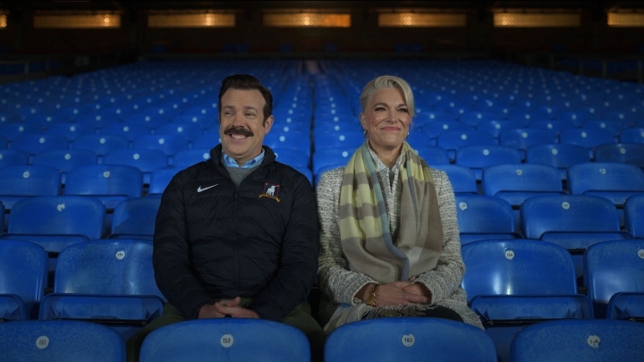 Ted und Rebecca sitzen auf Stadionsitzen und lachen gemeinsam in einer Szene aus Ted Lasso.