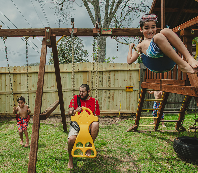 Mr. Muñoz sits on a backyard swing with his portable oxygen tank. His two sons, wearing swim trunks, swing enthusiastically next to him.