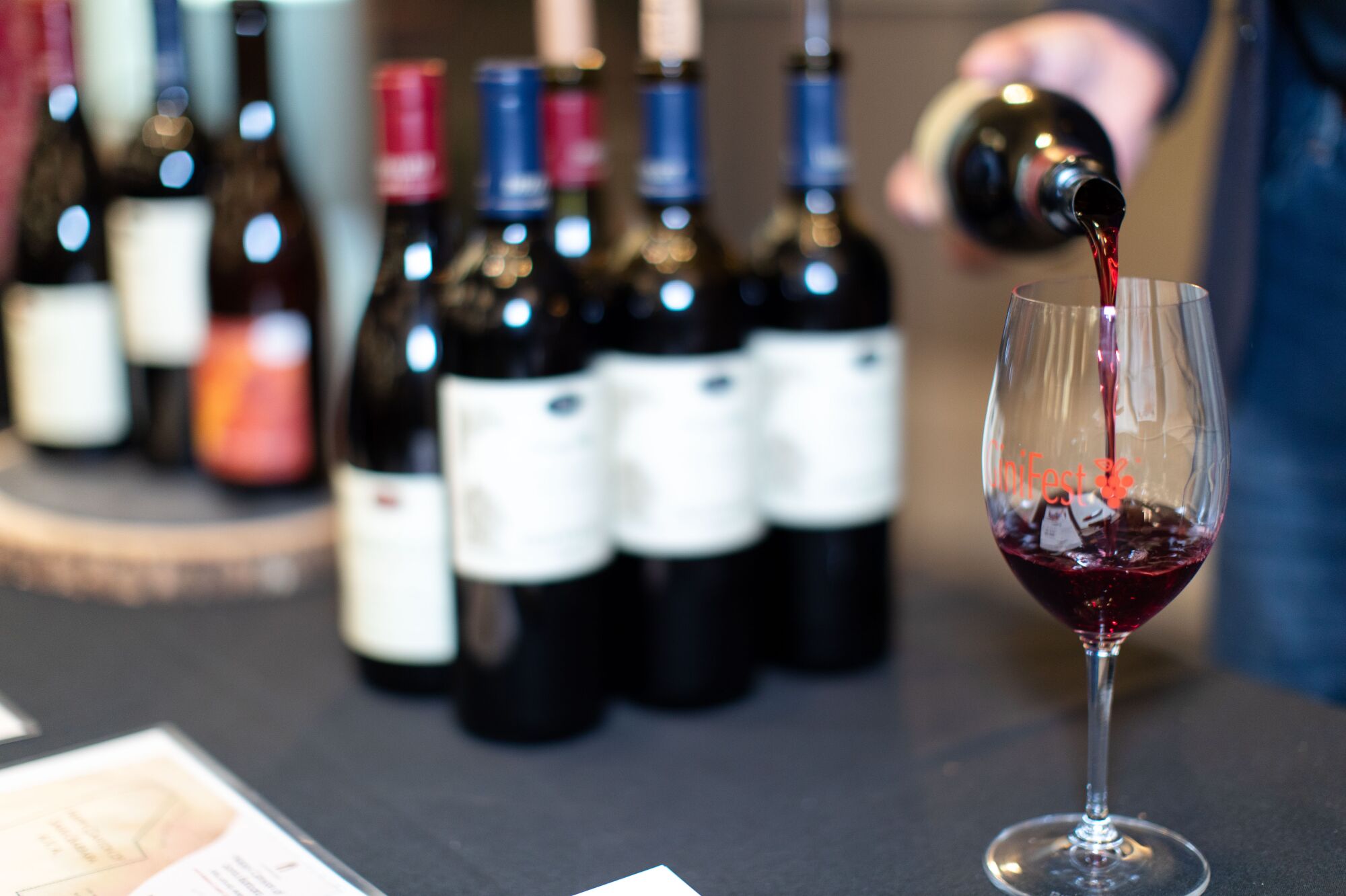 Eine Hand gießt Wein in ein Glas auf einer Theke, auf der mehrere Flaschen Wein stehen.