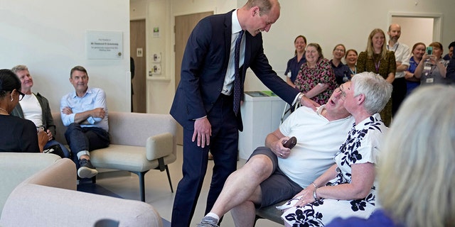 Prinz William in einem blauen Anzug lacht mit einem Mann in weißem Hemd und Shorts in einem Krebszentrum