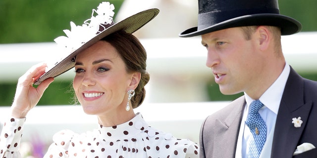 Kate Middleton trägt ein gepunktetes Kleid und einen passenden Hut und lächelt neben Prinz William in einem grauen Anzug und Zylinder