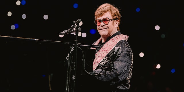 Elton John sitzt lächelnd am Klavier und trägt eine Sonnenbrille