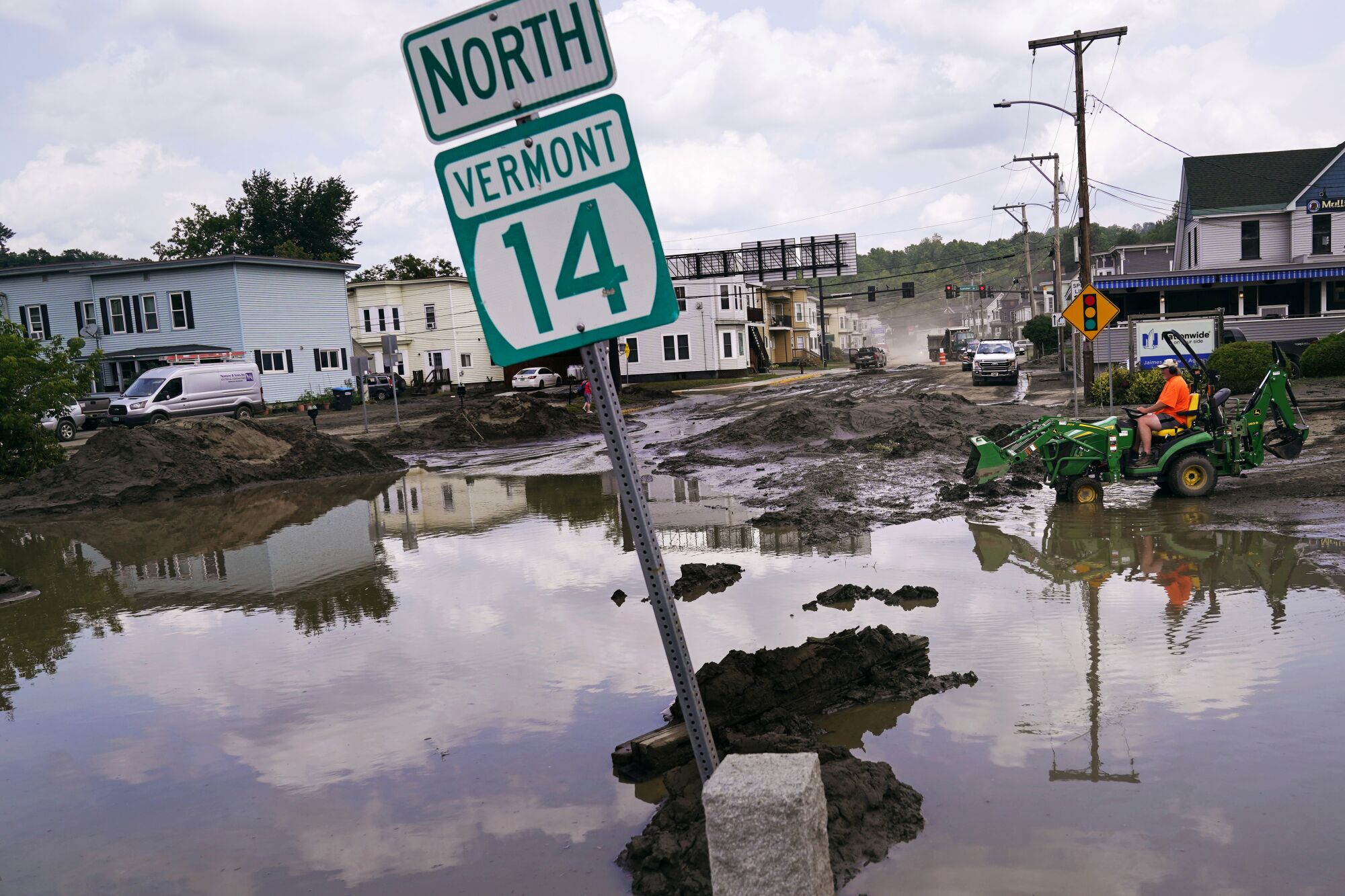 Ein kleiner Traktor fährt durch Schlamm neben Hochwasser, das teilweise ein Schild mit der Aufschrift überschwemmt "Nord-Vermont 14."
