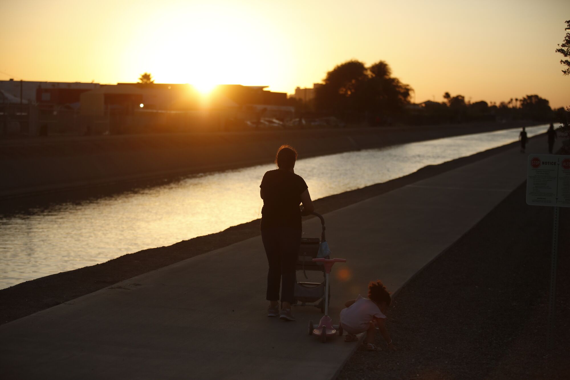Ein Kleinkind hockt und greift nach etwas abseits des Weges, während es einer Frau folgt, die bei Sonnenuntergang einen Wasserweg entlang geht