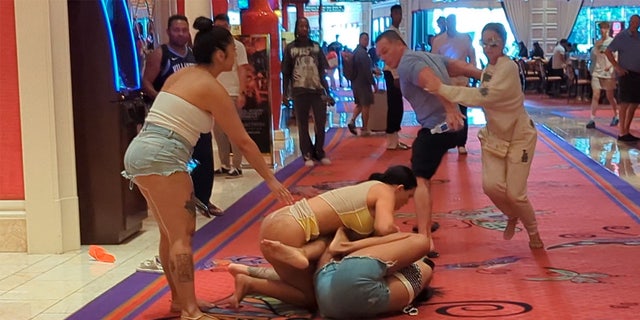 Eine Frau in einem Tanga wird gezeigt, wie sie mit einer Frau in abgeschnittenen Shorts auf dem Boden eines Casinos ringt.