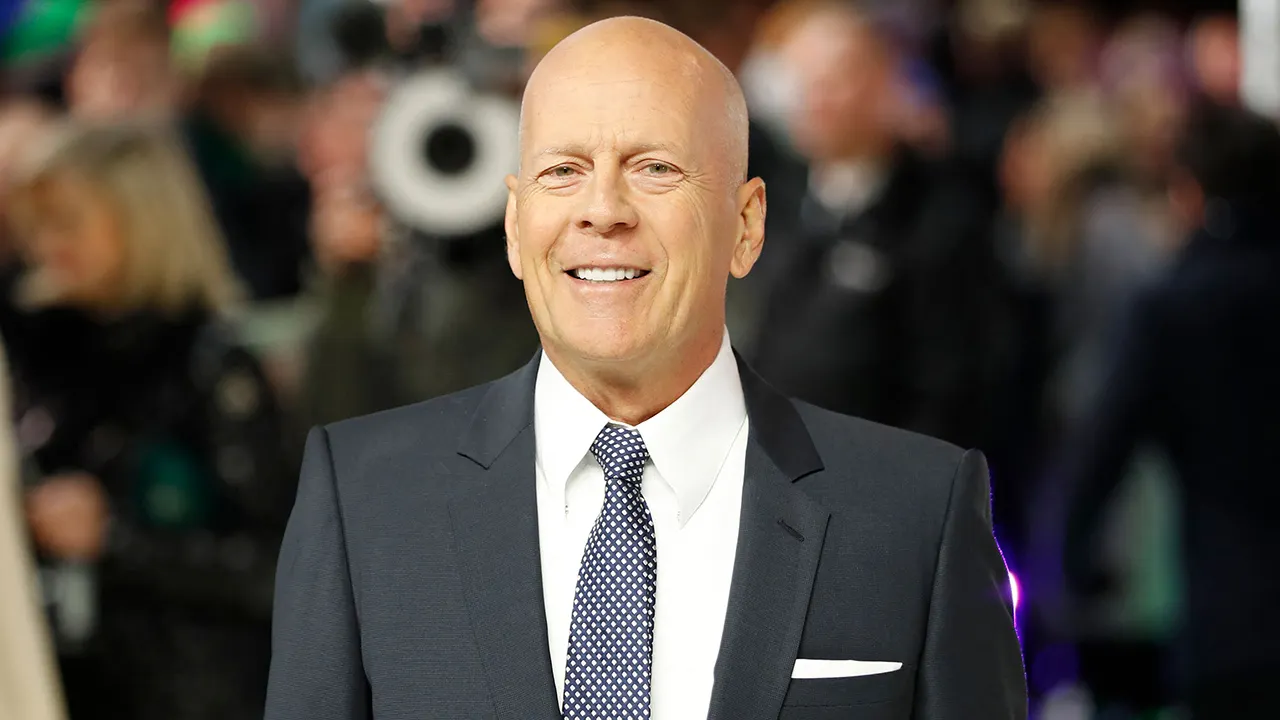 Bruce Willis lächelt auf dem roten Teppich und trägt einen schwarzen Anzug, weiße Knöpfe und eine blau gemusterte Krawatte