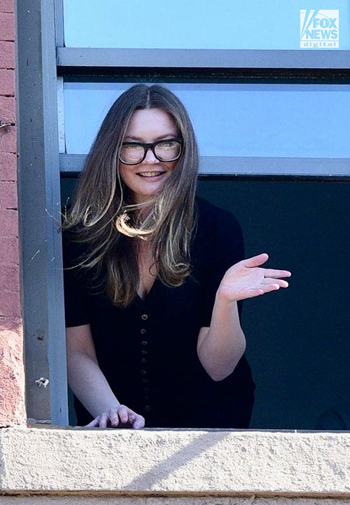 Anna Sorokin in schwarzem Top und Brille winkt aus dem Fenster eines Backsteingebäudes