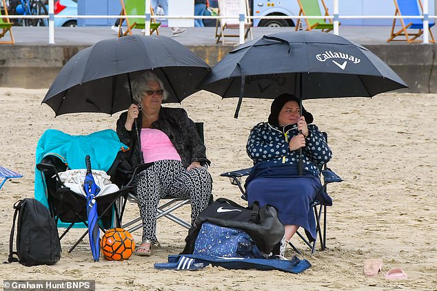 Holidaymakers on the beach sheltering under umbrellas in the rain at the seaside resort of Weymouth in Dorset on a day of overcast cloudy skies and light showers