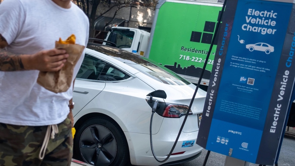 Ein Mann in Shorts und T-Shirt, der einen Burrito isst, geht an einem weißen Tesla vorbei, der an eine öffentliche Ladestation angeschlossen ist