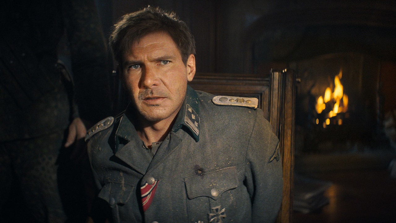 A still frame of Harrison Ford as Indiana Jones in a dirty blue jacket looking up and out ominously, his face is altered by AI