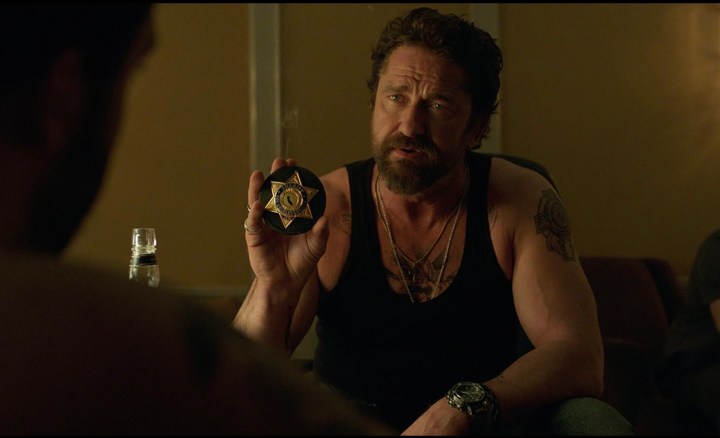 Gerard Butler sits down and holds up a police badge in Den of Thieves.