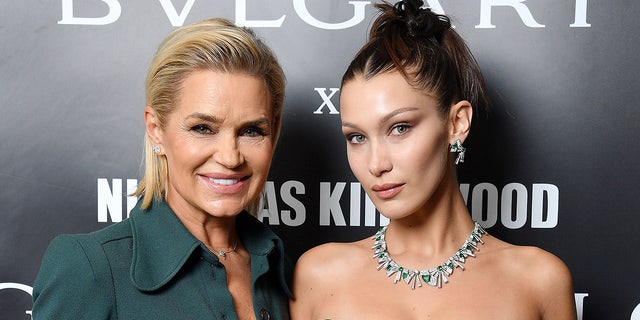 Yolanda Hadid in a dark teal blouse smiles next to daughter Bella Hadid in an up-do and black dress with a diamond necklace