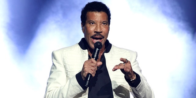 Lionel Richie tritt in weißer Jacke und schwarzem Hemd darunter auf der Bühne vor blau/weißem Hintergrund auf