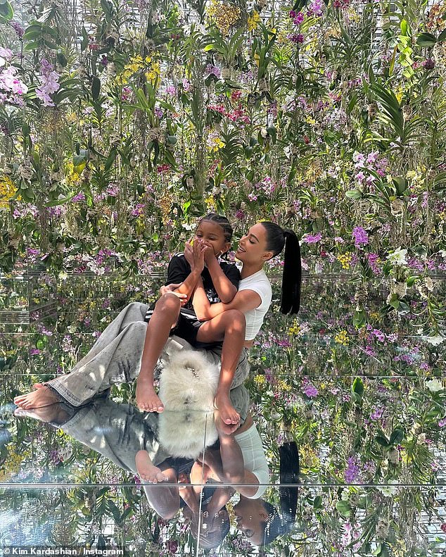 Schwebend: Kim sitzt mit Saint auf ihrem Schoß auf dem Boden, während der Boden und die Wände um sie herum tropisches Grün mit rosa Blumen reflektieren