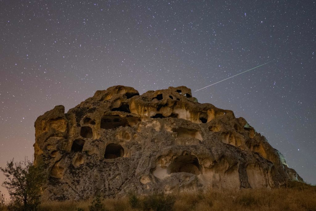 Perseiden-Meteoriten ziehen über einer großen Felsstruktur mit zahlreichen geschnitzten Höhlen und Öffnungen über den Himmel.