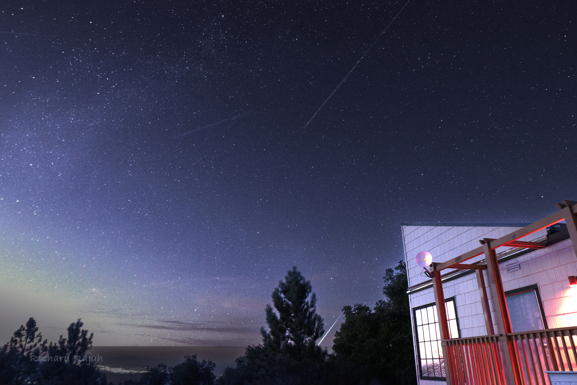 Perseiden-Meteore ziehen über den Himmel und im unteren Teil des Bildes ist ein heller Feuerball zu sehen, der sich direkt hinter den Bäumen bewegt.  Rechts im Bild ist ein Gebäude beleuchtet.
