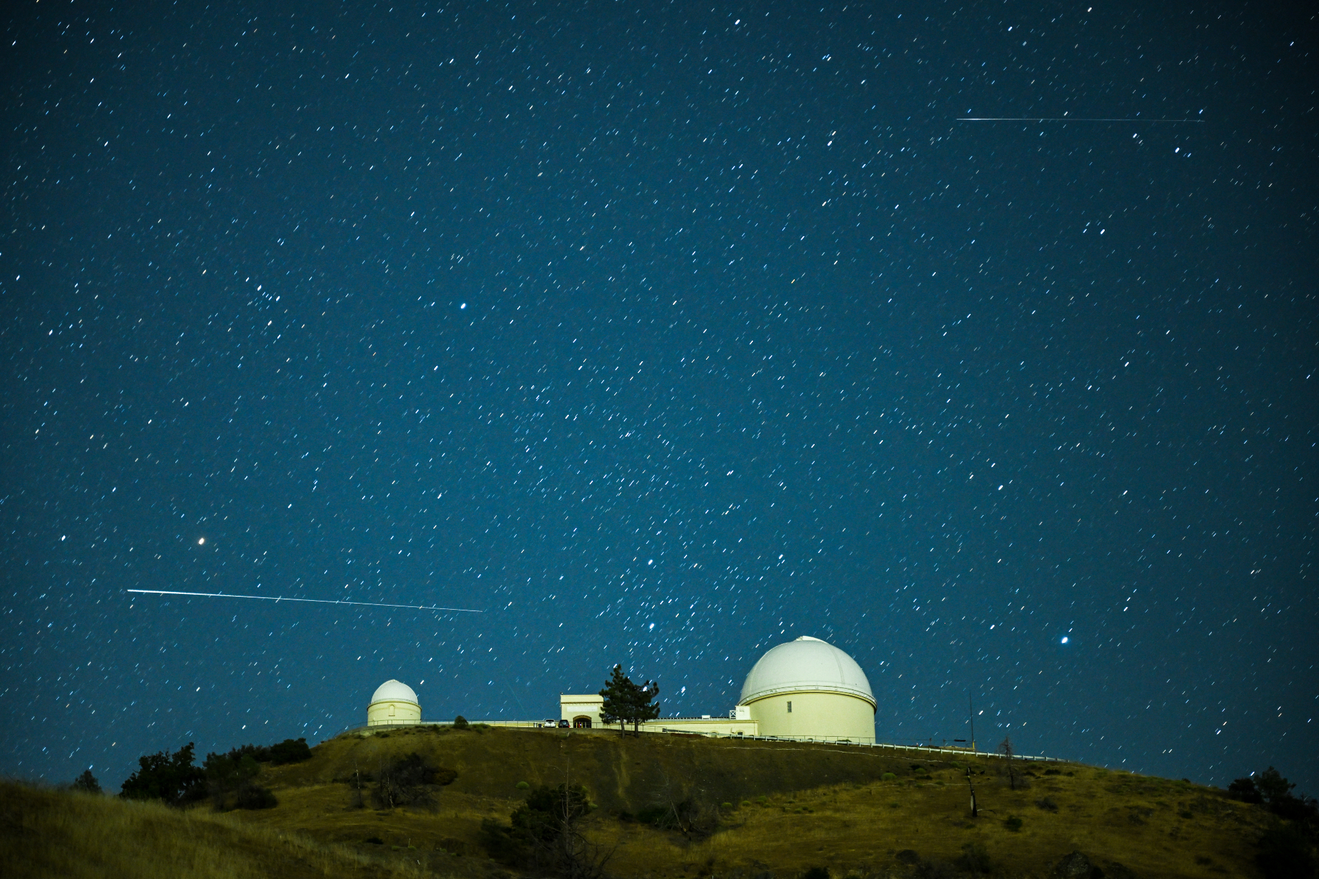 Ein langer weißer Zug eines Perseiden-Meteors fährt in horizontaler Ausrichtung vor einem Sternenhimmel über die Kuppelstruktur des Lick-Observatoriums.