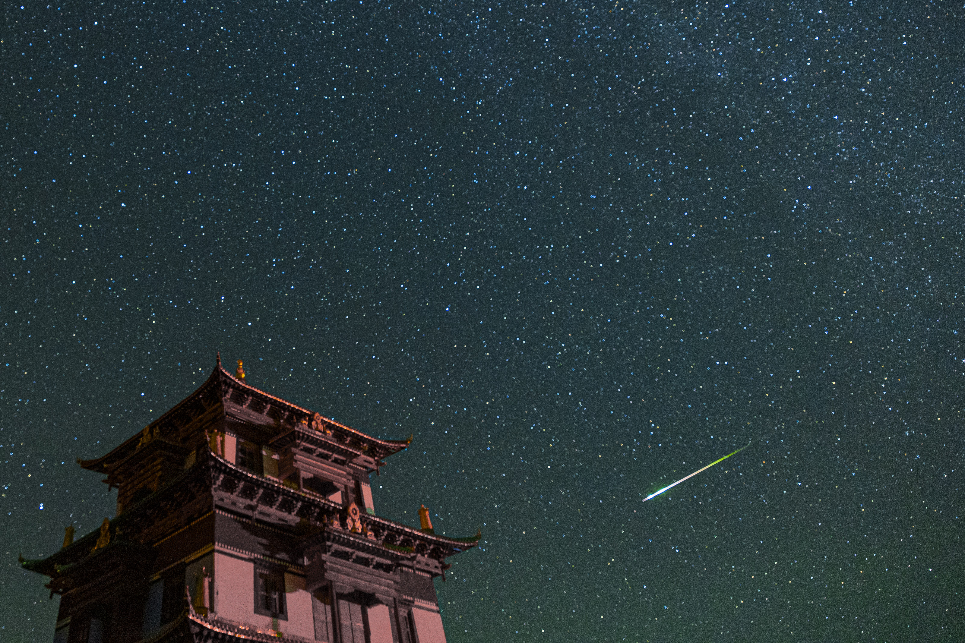 Ein großes Gebäude auf der linken Seite und ein leuchtend grün-weißer Perseiden-Meteor, der über den sternenübersäten Himmel streift.