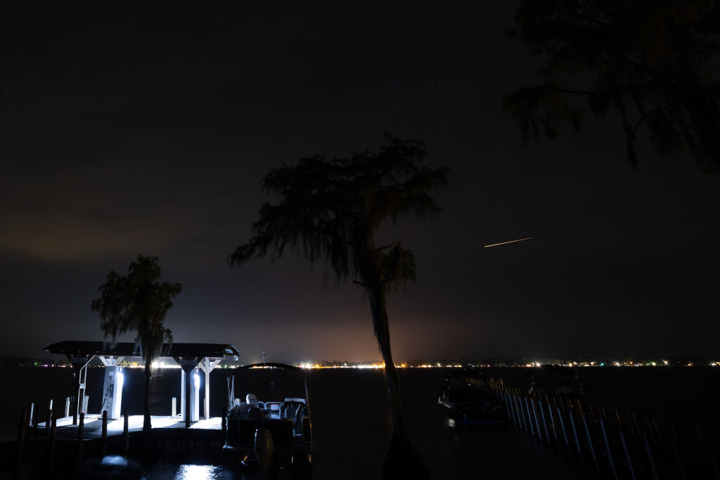 Ein perseidischer Meteor, der über helle Lichter neben einem großen See hinwegfliegt.