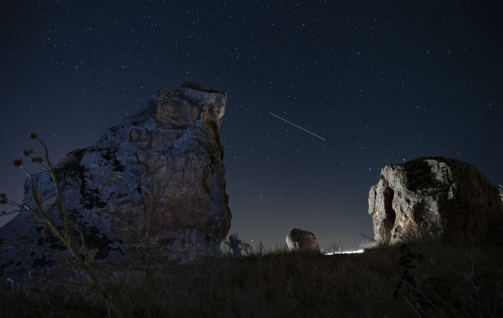 Ein langer Perseiden-Meteorzug zieht zwischen zwei großen Felsstrukturen im Vordergrund des Bildes über den Himmel.