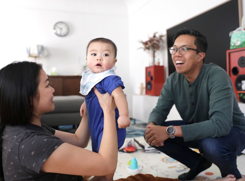 Ein Paar spielt mit einem Baby in einem Wohnzimmer.  Ein Elternteil hebt das Kind hoch, während das Kind in die Kamera schaut