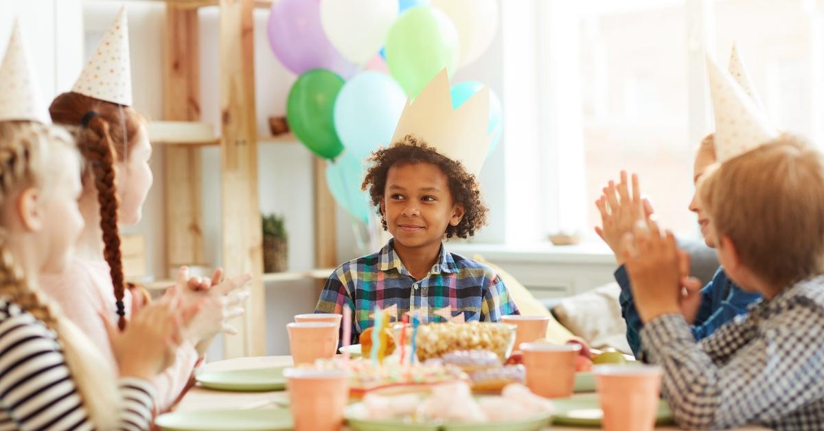 Kleiner Junge in kariertem Button-Down-Hemd und Geburtstagshut sieht schelmisch aus, während er mit seinen Freunden an einem dekorierten Tisch sitzt. 