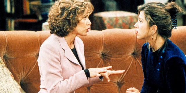 Jennifer Gray in einem rosa Blazer spielt Mindy Hunter und spricht mit ausgestreckten Händen mit Jennifer Aniston als Rachel Green in einer Folge von "Freunde" sitzt auf der orangefarbenen Couch