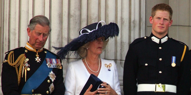 Prinz Harry in einem Militäranzug sieht genervt aus, während Camilla ein weißes Kleid und einen übergroßen marineblauen Hut trägt und neben Prinz Charles in einer Militäruniform steht