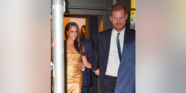 Meghan Markle trägt ein goldenes Kleid und wird von Prinz Harry in einem dunkelblauen Anzug und einem weißen Hemd begleitet