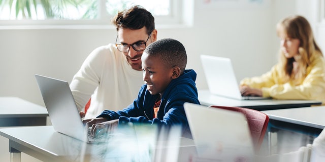 Ein männlicher Lehrer hilft einem kleinen Jungen beim computergestützten Lernen im Klassenzimmer.
