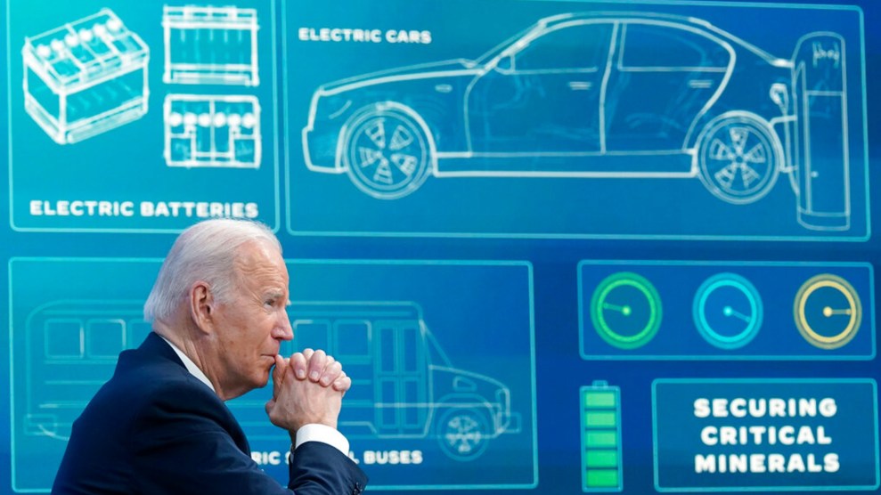 Joe Biden sitzt mit dem Kinn in gefalteten Händen vor einer blauen Tafel, auf der Informationen über Elektrofahrzeuge projiziert werden, darunter ein Umriss eines Autos und einer Batterie sowie die Worte "Sicherung kritischer Mineralien."