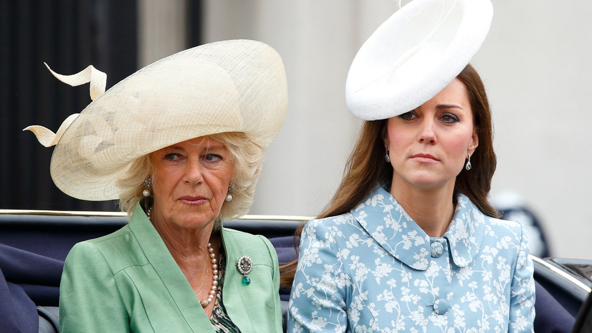 Königin Camilla trägt ein grünes Kleid und einen elfenbeinfarbenen Hut und sitzt neben Kate Middleton in einem blau-weißen Blumenkleid und einem weißen Hut
