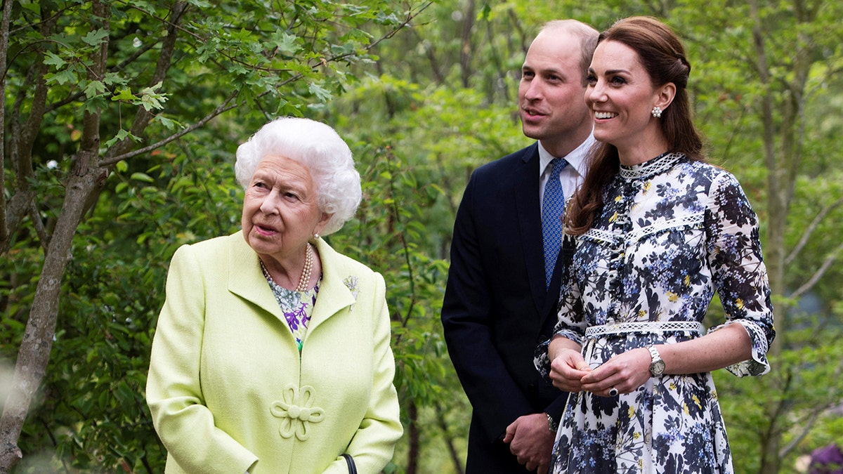 Königin Elizabeth trägt ein neongrünes Kleid, neben Kate Middleton in einem mehrfarbigen Kleid und Prinz William in einem dunkelblauen Anzug und Krawatte