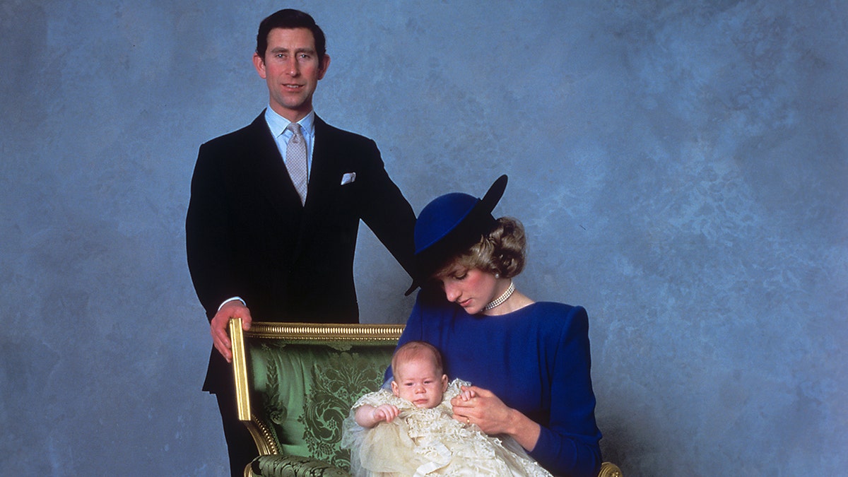 Prinzessin Diana trägt ein blaues Kleid und einen Hut und hält Prinz Harry, während Prinz Charles im Anzug in die Kamera blickt